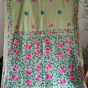May include: A pale green textile with detailed floral embroidery. The design incorporates pink flowers and teal leaves, with a matching border. The fabric is displayed hanging, highlighting the craftsmanship and colour scheme.
