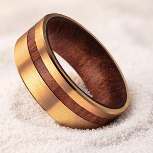 Puede incluir: Un anillo con un exterior de metal en tono dorado cepillado y una incrustación central de madera de color marrón oscuro. La banda tiene un perfil liso y redondeado con un acabado pulido en los bordes internos, resaltando el contraste de texturas.