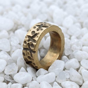 May include: A gold-toned metal ring with a textured, matte finish. The outer surface of the band features a repeating pattern of recessed circular indentations and cross-shaped markings, creating a rugged, uneven appearance around the circumference.