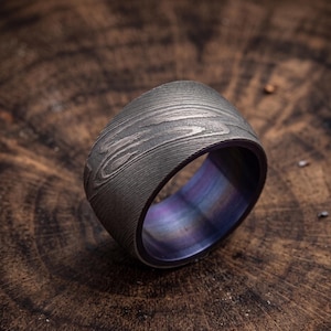 May include: A wide, textured ring with a wood grain pattern in shades of gray and silver. The interior of the ring has a purple and blue iridescent finish. The ring is set against a dark, wooden background.