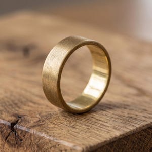 Puede incluir: Un anillo de latón cepillado con un perfil plano. El anillo es de color dorado cálido y se encuentra sobre una superficie de madera. El diseño sencillo del anillo lo hace adecuado para el uso diario.