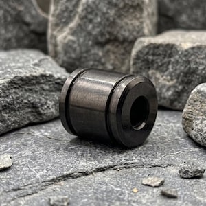May include: A cylindrical metal bead with a dark, gunmetal gray finish and a smooth, polished surface. The bead features a central horizontal groove encircling its body and a circular opening running through the middle of the component.