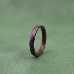Può includere: Primo piano di un anello con un motivo unico e vorticoso di colori. L'anello presenta sfumature di marrone, blu e arancione, creando un effetto marmorizzato. L'anello è posto su uno sfondo verde tenue.