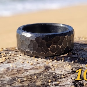 Op de afbeelding: Een zwarte ring met gehamerde textuur. De ring heeft een brede band en een getextureerd oppervlak. De ring is maat 10. De ring wordt getoond op een stuk drijfhout met een vage strandachtergrond.