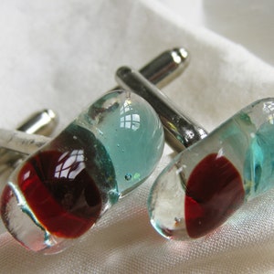 May include: A pair of cufflinks with a silver metal base and glass insets. The glass is clear with red and blue swirls.