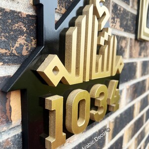 May include: A black and gold house-shaped sign with the Arabic word for "Allah" in gold lettering and the number "1034" in gold lettering. The sign is mounted on a brick wall. The text "Islamic Arabic Decor&reg;" is at the bottom of the sign.