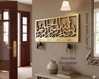 Op de afbeelding: Een goudkleurige metalen sleutelhouder met Arabische kalligrafie. De sleutelhouder heeft vier haken en de woorden "Bismillah Keyholder" staan eronder. Sleutels hangen aan de haken. Het is bevestigd aan een beige muur.