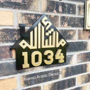 May include: Black house-shaped sign with gold Arabic calligraphy and the number 1034. The sign is mounted on a brick wall. The text "Islamic Arabic Decor" is at the bottom of the sign.