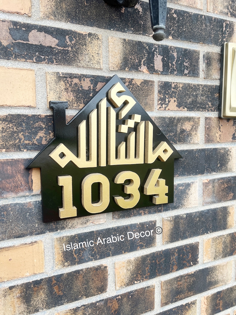 May include: Black house-shaped sign with gold Arabic calligraphy and the number 1034. The sign is mounted on a brick wall. The text "Islamic Arabic Decor" is at the bottom of the sign.