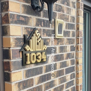 May include: A gold house-shaped sign with Arabic calligraphy and the number 1034. The sign is mounted on a brick wall with a black light fixture above it.