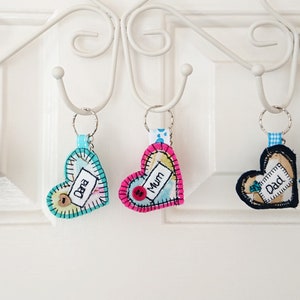 May include: Five heart-shaped and one star-shaped keyrings with blue, pink, and green fabric. Each keyring has a white tag with a name written on it:  "Csaba", "Dana", "Mum", "Dad", and "Daddy".