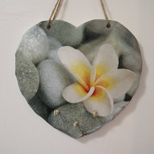 May include: A heart-shaped slate key holder with a floral design. The holder features a white flower with a yellow center and gray stones. There are three gold hooks on the bottom of the holder.