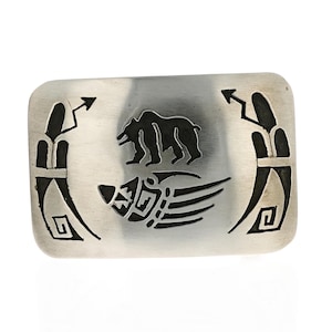 May include: A silver belt buckle featuring a black bear and paw print design. The buckle is decorated with intricate geometric patterns.