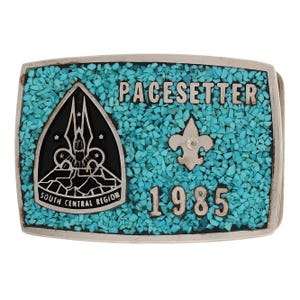May include: Silver-toned belt buckle with turquoise inlay. The buckle features the word "PACESETTER" and the year "1985". A black and silver emblem and a fleur-de-lis are also present. The buckle is rectangular with rounded corners.
