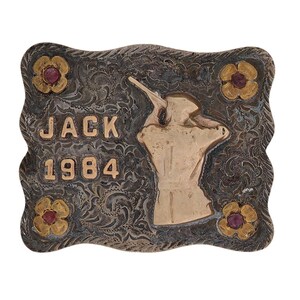 May include: A rectangular silver-toned belt buckle with a gold-toned figure of a person aiming a rifle. The buckle has the name "JACK" and the year "1984" in gold lettering. Four gold-toned floral accents with red centers are placed around the edges.