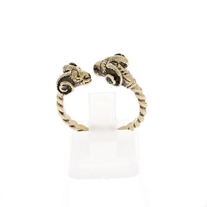 May include: A bronze ring with two ram heads facing each other. The ring has a twisted band design. The ram heads have detailed horns and facial features. The ring is displayed on a white stand against a white background.