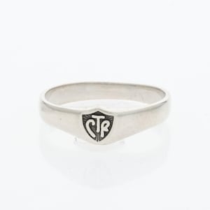 May include: A polished silver-toned ring featuring a central shield-shaped emblem. The emblem contains the letters CTR in a stylized black font. The band is smooth and uniform in width, with a metallic finish that reflects light across its surface.