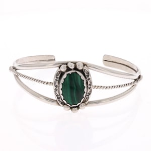 May include: Silver cuff bracelet with a deep green malachite stone. The oval stone is set in a scalloped bezel, surrounded by small silver beads. The bracelet has a three-strand design with a twisted rope detail.
