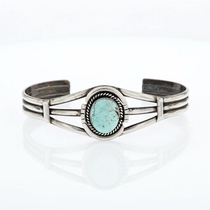 May include: A silver cuff bracelet featuring a central oval turquoise stone with a rope-textured border. The band is constructed from three parallel silver wires that converge at the sides, showcasing a polished metallic finish.