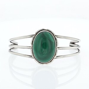 May include: A silver cuff bracelet featuring a central oval malachite stone with deep green swirling patterns. The stone is set in a serrated silver bezel, and the band consists of three parallel silver wires that meet at the sides.