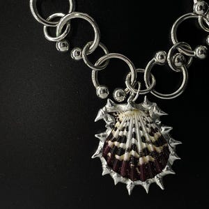 May include: A silver-toned necklace featuring a large, spiky pendant encasing a seashell with black, white, and maroon patterns. The necklace has a chunky chain design with large, circular links and smaller ball accents, creating a bold, eye-catching accessory.