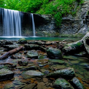 May include: A cascading waterfall flows over moss-covered rocks into a clear pool of water. A large, weathered tree trunk lies in the foreground, partially submerged in the water.