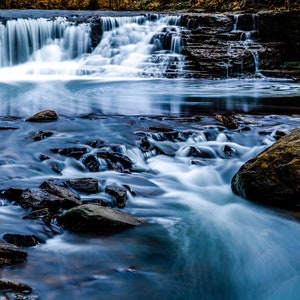 May include: A close-up of a waterfall cascading over rocks, with the water flowing smoothly over the rocks in the foreground. The water is a light blue color, and the rocks are a dark brown color.