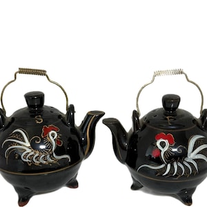 May include: Two black ceramic salt and pepper shakers shaped like miniature teapots. Each features a hand-painted rooster with red, white, and gold details. The left shaker is marked with an S and the right with a P. Both have metal wire handles.