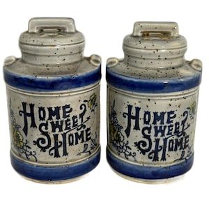May include: A pair of ceramic canisters with a speckled gray finish, blue accents, and the words "HOME SWEET HOME" in dark blue. Floral and leaf designs in yellow and blue decorate the sides. Each canister has a handle on top.