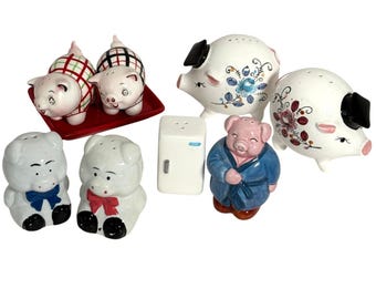 May include: A collection of ceramic salt and pepper shakers featuring various pig designs, including two on a red tray, two with coloured bows, two with floral patterns and black ears, one shaped like a refrigerator, and one wearing a blue robe.