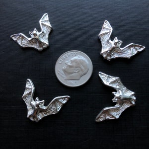 May include: Five silver-colored bat charms, each with a detailed design, are arranged on a black surface. A silver US dime coin is included for size comparison.