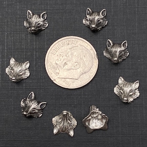 Op de afbeelding: Een verzameling van negen zilveren vossenkopcharms, gerangschikt in een cirkel rond een Amerikaanse dime. De charms zijn gedetailleerd en hebben een textuur.