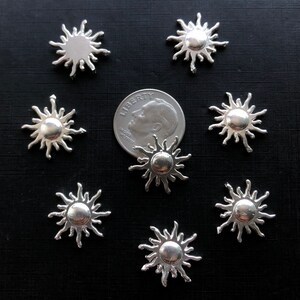 May include: Eight silver sun charms with a raised center, arranged on a black surface. The charms are approximately the size of a US quarter.
