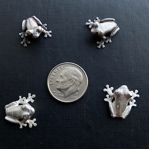 May include: Four silver frog figurines, each about the size of a UK ten pence coin, on a dark grey surface.