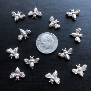 May include: A collection of 12 silver bee charms arranged in a circle around a US dime. The bee charms are about the size of a dime.