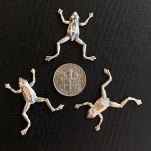 May include: Three silver frog figurines, each with a different pose, are arranged on a black surface. A UK ten pence coin is placed in the centre of the image for scale.