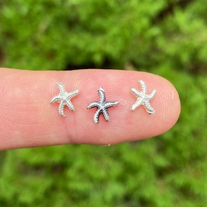 May include: Three silver starfish charms, each with a slightly different design, are displayed on a finger.