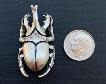 May include: Silver-toned metal scarab beetle pendant with detailed legs, head, and wing details. A U.S. dime is next to the pendant for size comparison. The pendant is on a black surface.
