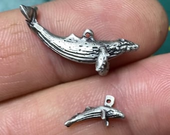 May include: Two silver whale charms, one larger than the other. The larger whale charm is in a swimming position with its tail up. The smaller whale charm is in a similar position but is smaller in size.
