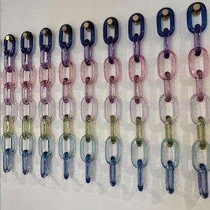 以下が含まれることがあります： 青、ピンク、紫、緑など、さまざまな色のガラス製の装飾チェーンリンクが9つ、白い壁に取り付けられています。各チェーンは、金色のスタッドで壁に取り付けられています。