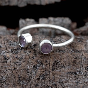 Könnte beinhalten: Ein silberner Ring mit zwei runden, hellvioletten Edelsteinen. Der Ring hat ein offenes Design, wobei die Edelsteine an gegenüberliegenden Enden des Bandes angebracht sind. Der Ring wird auf einer strukturierten, braunen Holzoberfläche präsentiert.