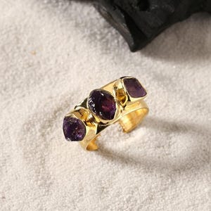 Könnte beinhalten: Ein goldfarbener Armreifring mit drei Amethyststeinen. Der Ring hat eine polierte Oberfläche und ist vor einem sandigen Hintergrund platziert. Die Amethyststeine sind tiefviolett.