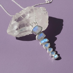 May include: A silver necklace featuring a pendant with seven blue and white moonstone pieces. The pendant is set in a silver bezel and hangs from a delicate silver chain. The necklace is displayed on a clear crystal against a purple background.