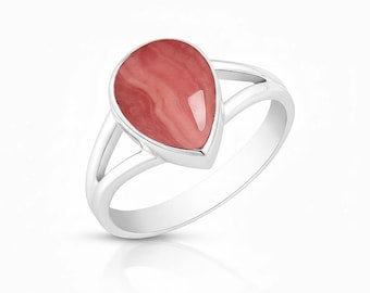 Puede incluir: Un anillo de plata con una piedra preciosa rosa en forma de pera con un aspecto veteado. La piedra preciosa está engastada en un bisel de plata y el anillo tiene un diseño de caña dividida. El anillo se muestra sobre un fondo blanco.