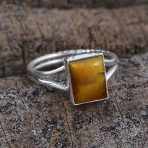 May include: A silver ring with a rectangular, golden-brown tiger's eye stone. The ring has a double band design, with the stone set in a silver bezel. The ring is displayed on a wooden surface.