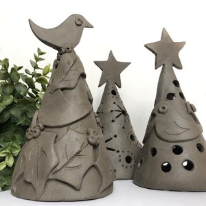 Op de afbeelding: Drie bruine kerstboomversieringen van keramiek. De boom links heeft een vogel bovenop en bladeren rond de basis. De middelste boom heeft een ster bovenop en is versierd met gaten. De boom rechts heeft een ster bovenop en een vogel aan de voorkant.