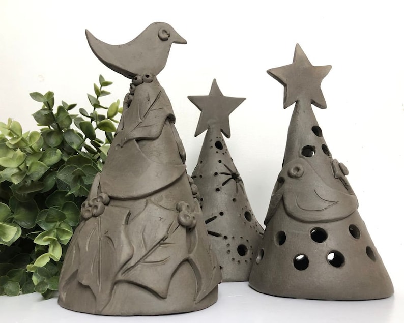 Op de afbeelding: Drie bruine kerstboomversieringen van keramiek. De boom links heeft een vogel bovenop en bladeren rond de basis. De middelste boom heeft een ster bovenop en is versierd met gaten. De boom rechts heeft een ster bovenop en een vogel aan de voorkant.
