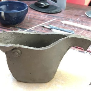 Peut inclure: Un pot en argile gris avec une anse et un petit trou rond sur le c&ocirc;t&eacute;. Le pot est inachev&eacute; et a une texture rugueuse.