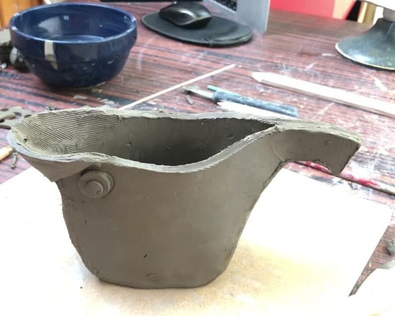 Peut inclure: Un pot en argile gris avec une anse et un petit trou rond sur le c&ocirc;t&eacute;. Le pot est inachev&eacute; et a une texture rugueuse.