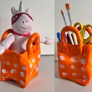 May include: Orange ceramic pencil holder with white polka dots shaped like a shopping bag. A pink unicorn plush toy is sitting inside the holder.